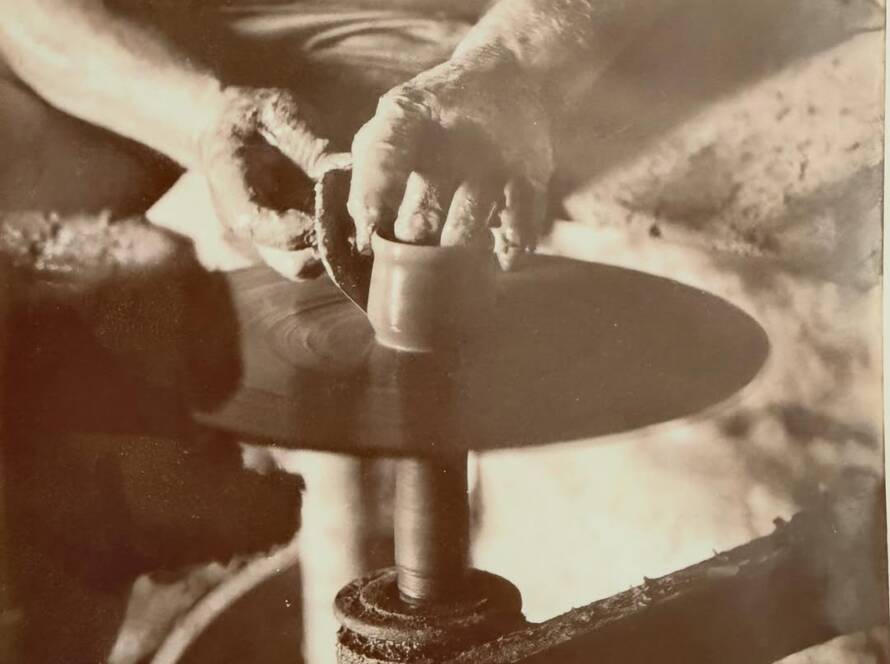 A vintage black-and-white photograph of a master potter’s hands shaping clay on a traditional wheel at the Thassos workshop.